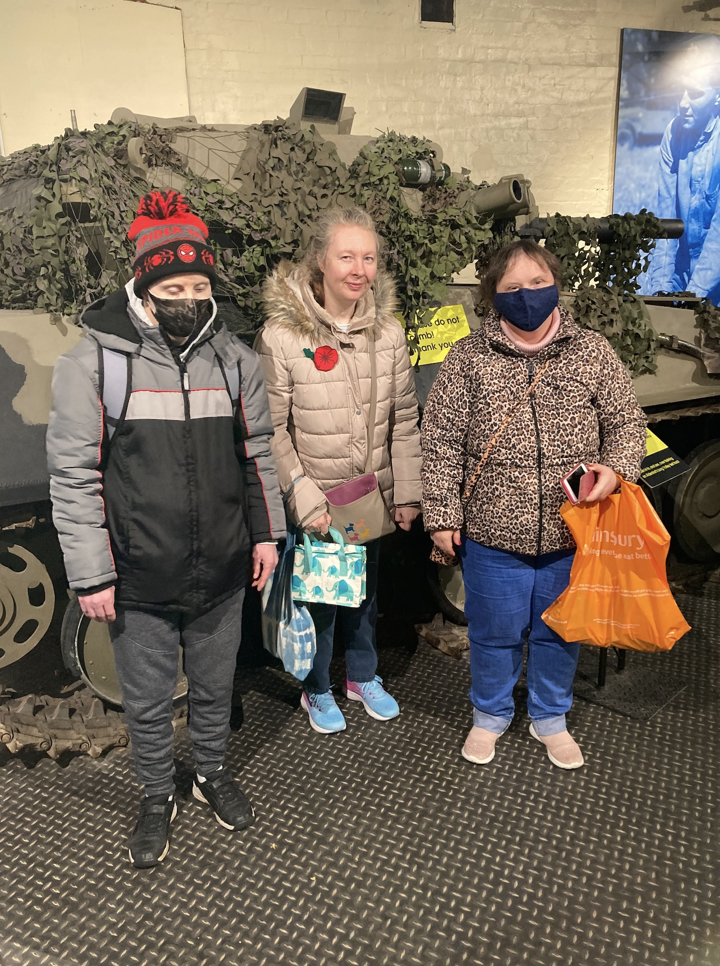 Some of the Farnham Group are stood in front of an army tank replica. They are smiling at the camera and two out of three people are wearing a face mask.
